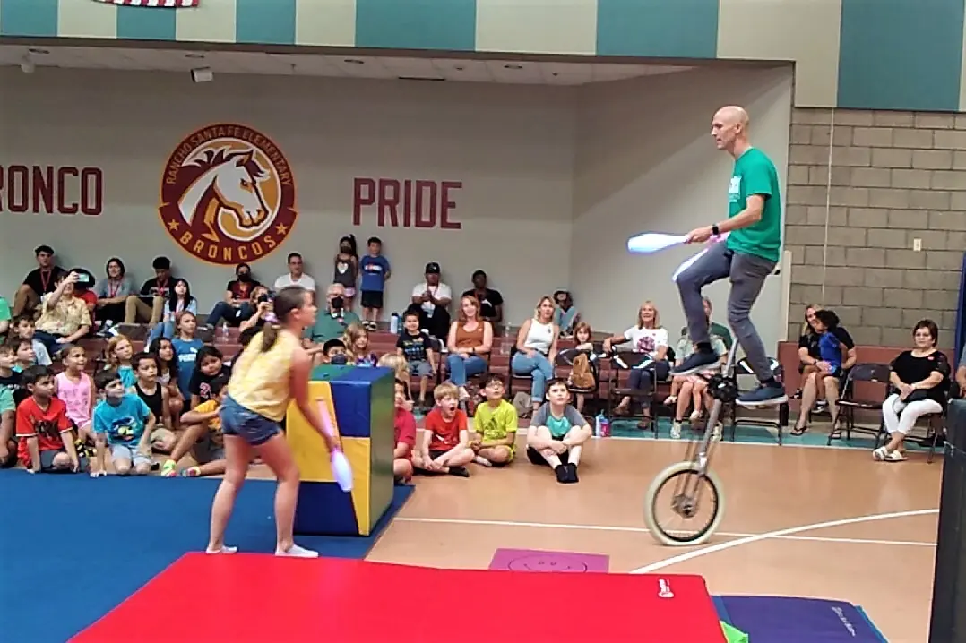 Dan juggling on a giraffe unicycle as a student tosses him a club during a school circus show