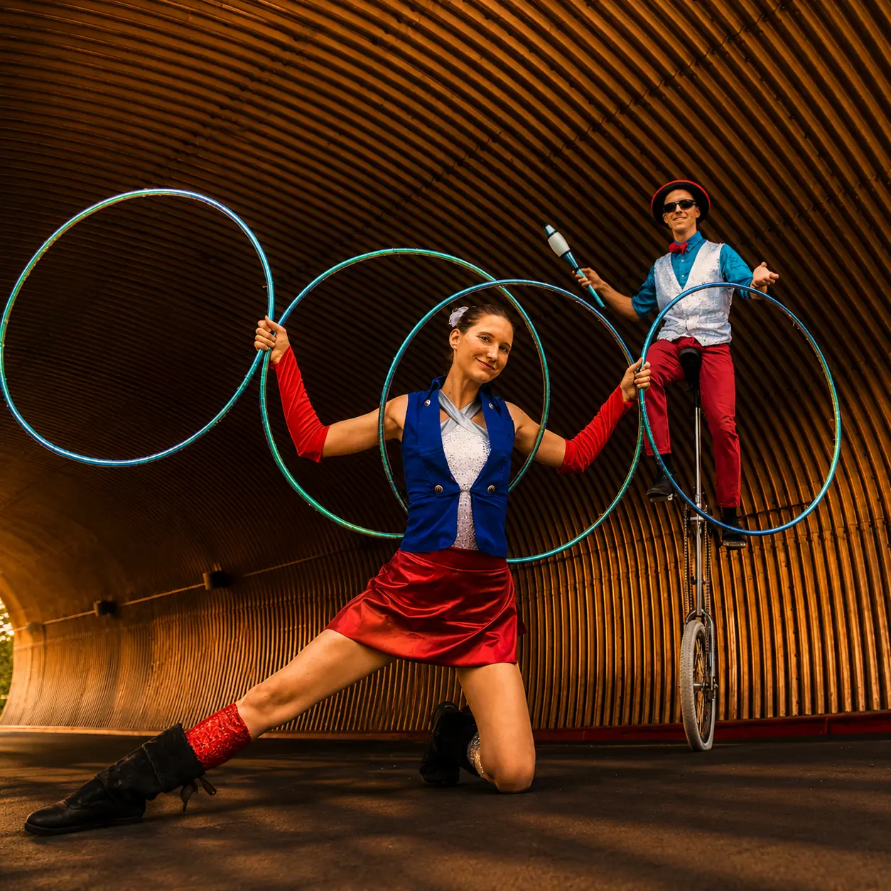 Tabitha Kelley posing with four hoops while Dan Barber balances on a giraffe unicycle